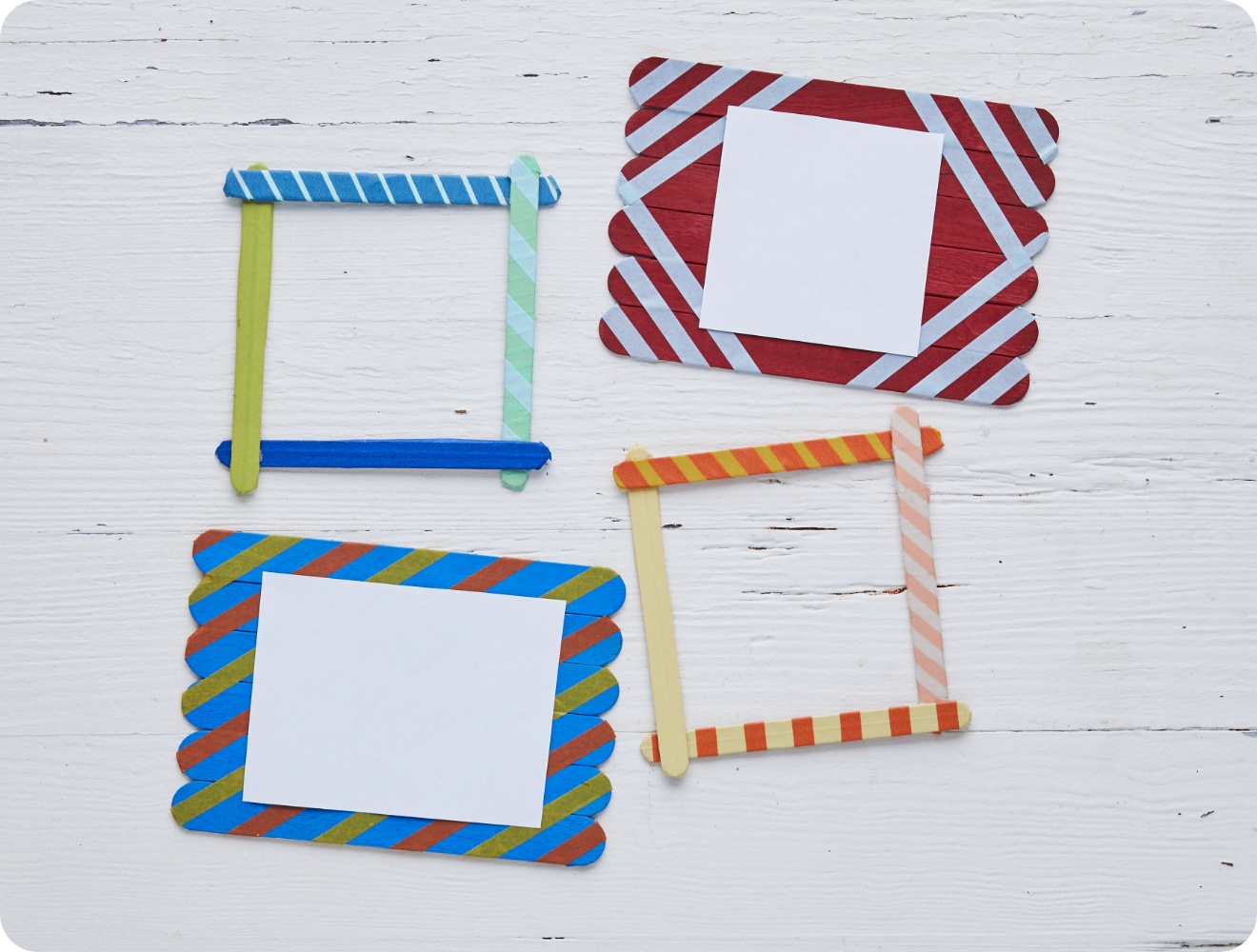 Bunte DIY-Bilderrahmen aus Eisstielen mit Washi-Tape-Dekor auf weißem Holztisch. Zwei Rahmen enthalten leere Papierstücke.