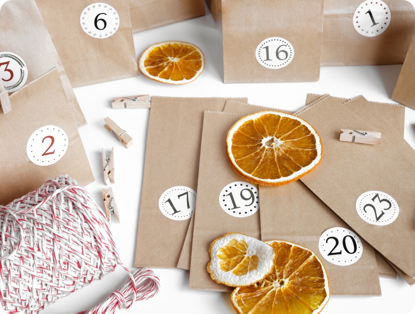 DIY-Adventskalender aus Papiertüten mit Zahlenstickern, Holzklammern, getrockneten Orangenscheiben und Bäckergarn auf weißem Hintergrund.