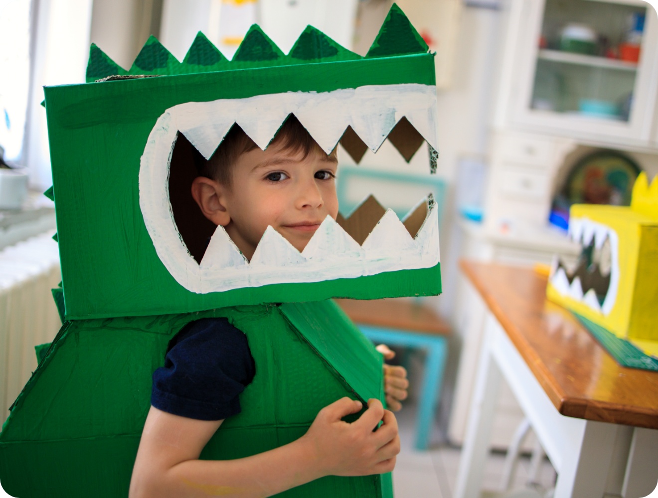 Lächelnder Junge trägt ein selbstgebasteltes Dinosaurier-Kostüm aus grün bemalten Kartons mit weißen Zähnen. Kreatives DIY-Projekt.
