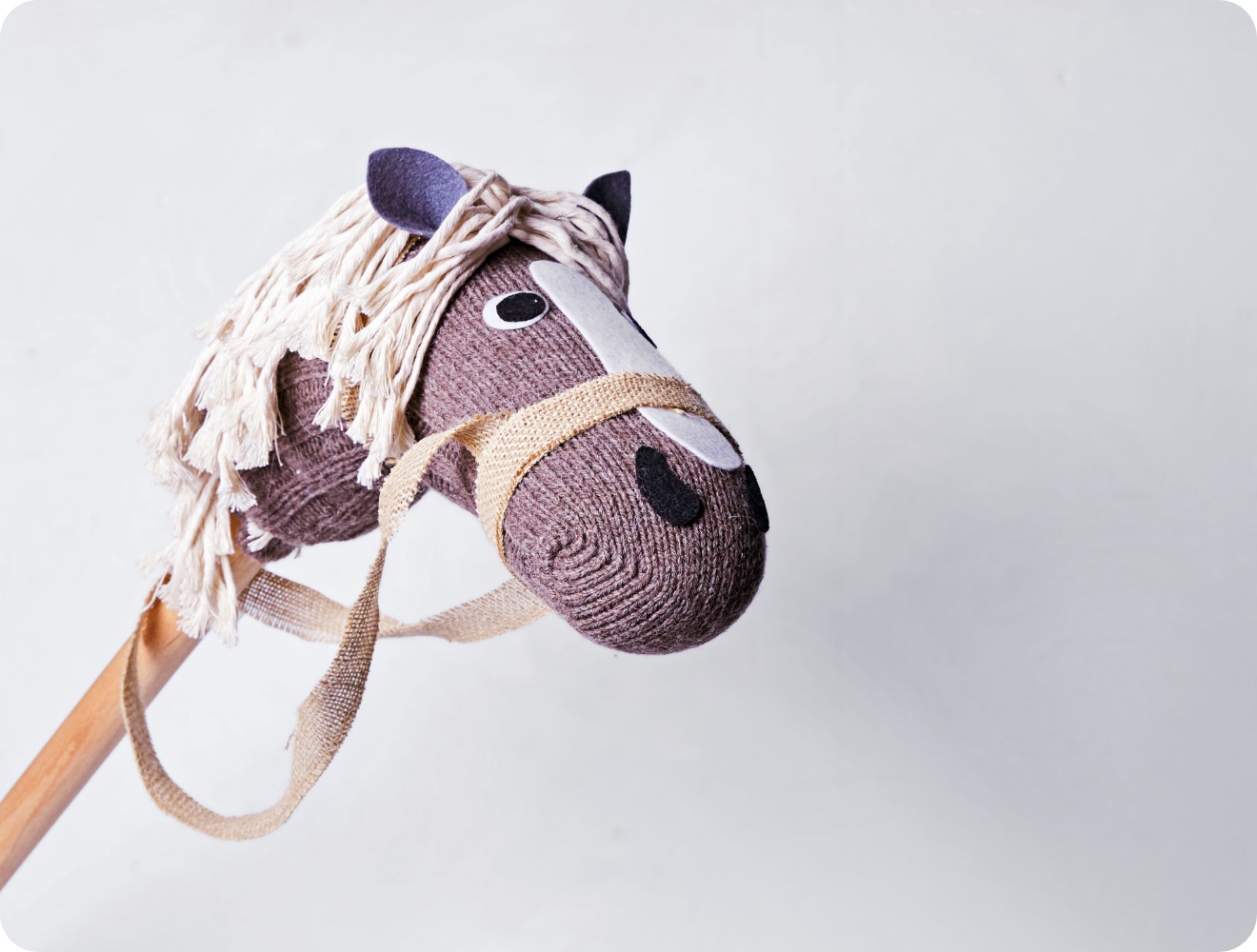 Selbstgemachtes Steckenpferd mit gestricktem Kopf, Jutegeschirr und heller Wollmähne vor neutralem Hintergrund. Kreatives DIY-Spielzeug.