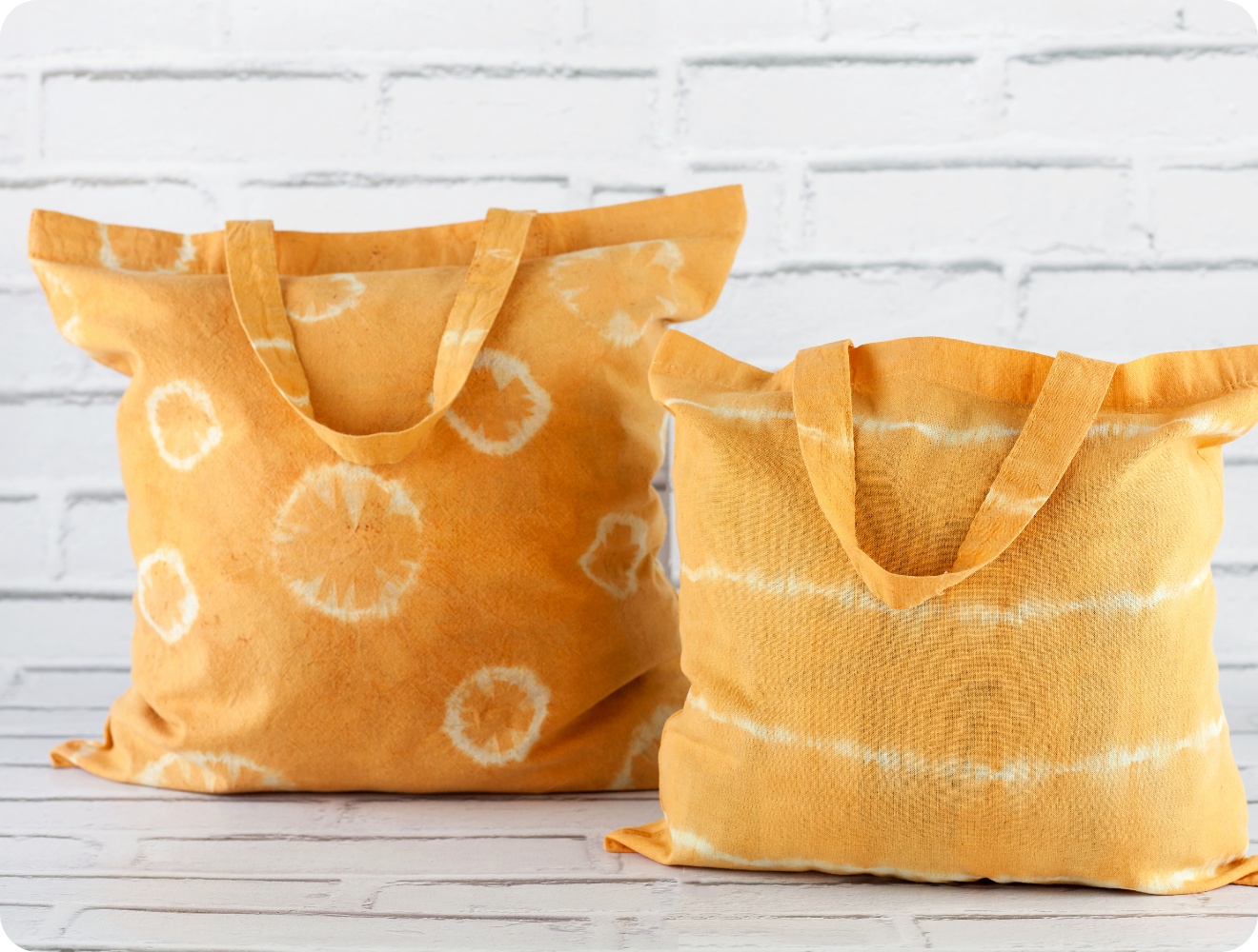 Zwei handgefärbte Stofftaschen in Gelb mit Batikmustern vor weißer Ziegelwand. Nachhaltige DIY-Taschen mit individuellem Design.