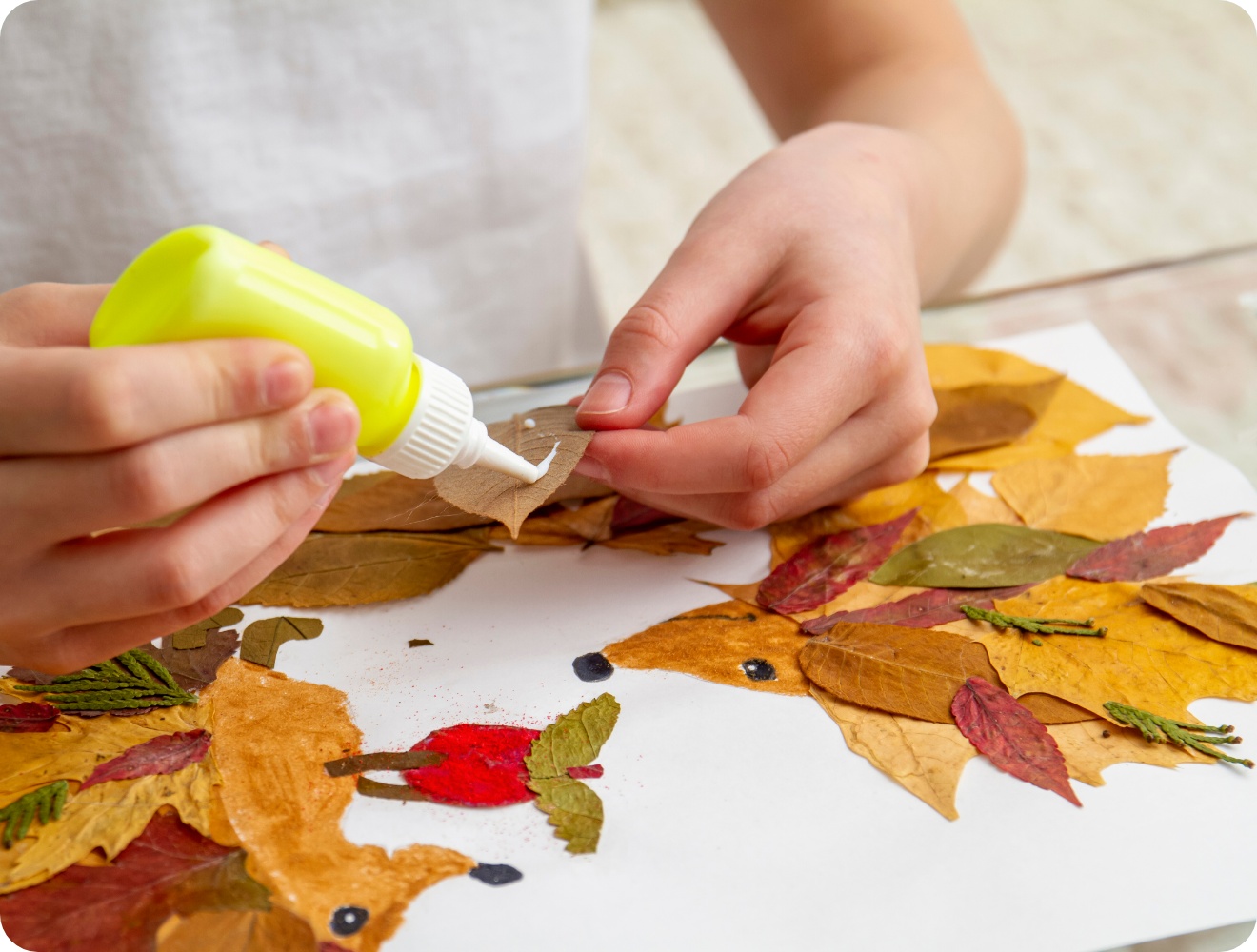 Kind bastelt ein herbstliches Bild mit getrockneten Blättern und Kleber. Kreatives DIY-Projekt mit Blätterkunst auf weißem Papier.