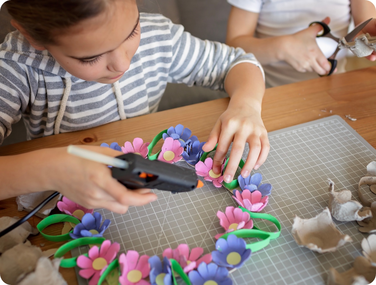 Mädchen bastelt mit Heißkleber einen bunten Blumenkranz aus recyceltem Eierkarton. Kreatives DIY-Projekt auf Holztisch.