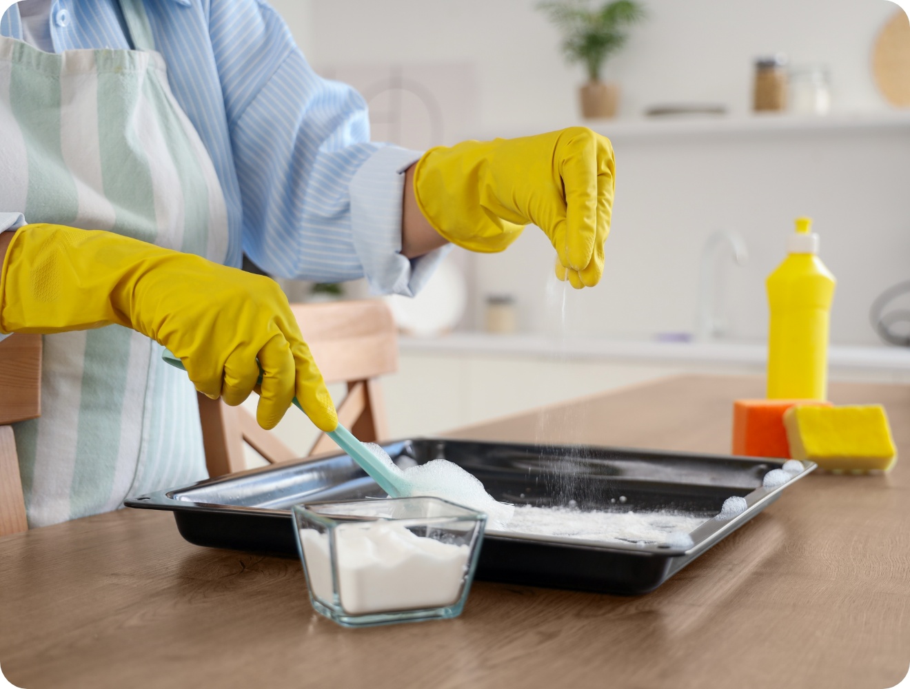 Person mit gelben Gummihandschuhen reinigt ein Backblech mit Natron und Wasser in einer modernen Küche.