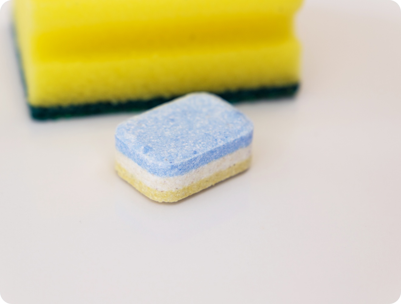 Nahaufnahme eines bunten Spülmaschinentabs vor einem gelben Schwamm auf einer weißen Oberfläche.