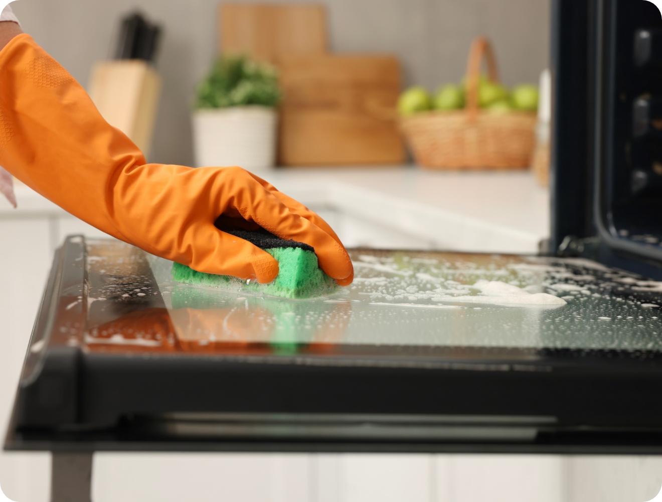 Person mit orangefarbenem Gummihandschuh reinigt die Glasinnenseite einer offenen Backofentür mit einem grünen Schwamm.
