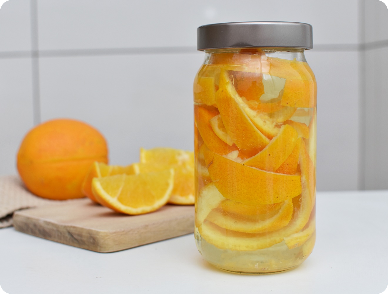 Glas mit Orangenschalen in Essig zur Herstellung eines natürlichen Reinigers, daneben frische Orangen auf einem Schneidebrett.
