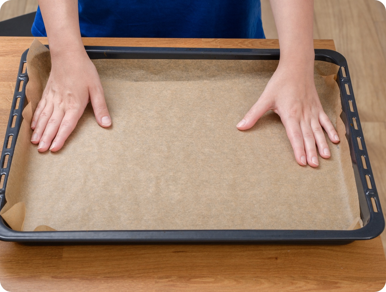 Person legt ein Stück Backpapier auf ein schwarzes Backblech, das auf einem Holztisch liegt.