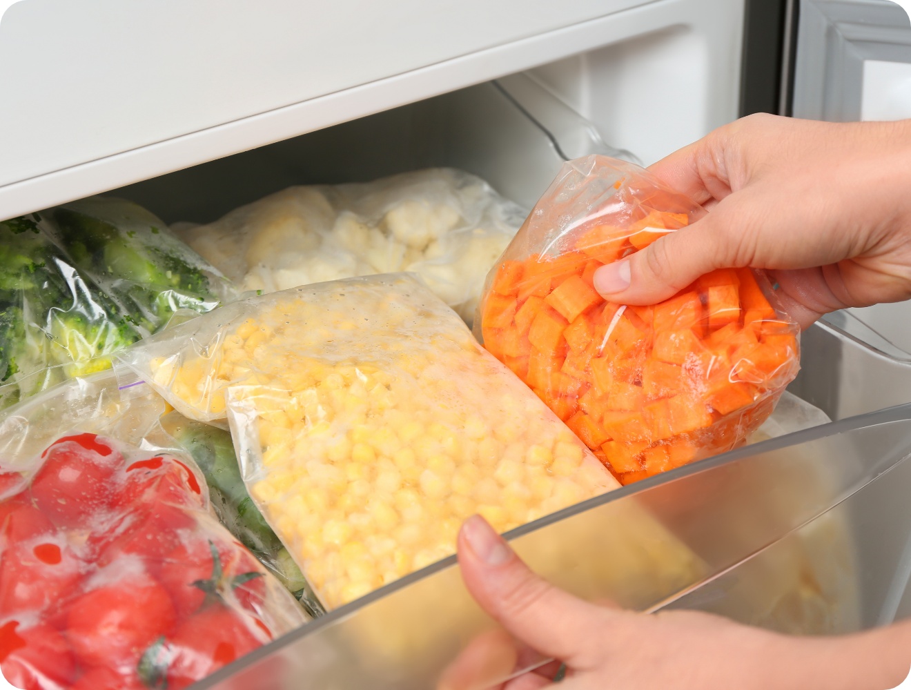 Person legt gefrorene und in einer Plastiktüte verpackte Möhrenwürfel in ein Gefrierfach. In der Schublade befinden sich außerdem abgepackter und gefrorener Mais, Brokkoli und Tomaten.