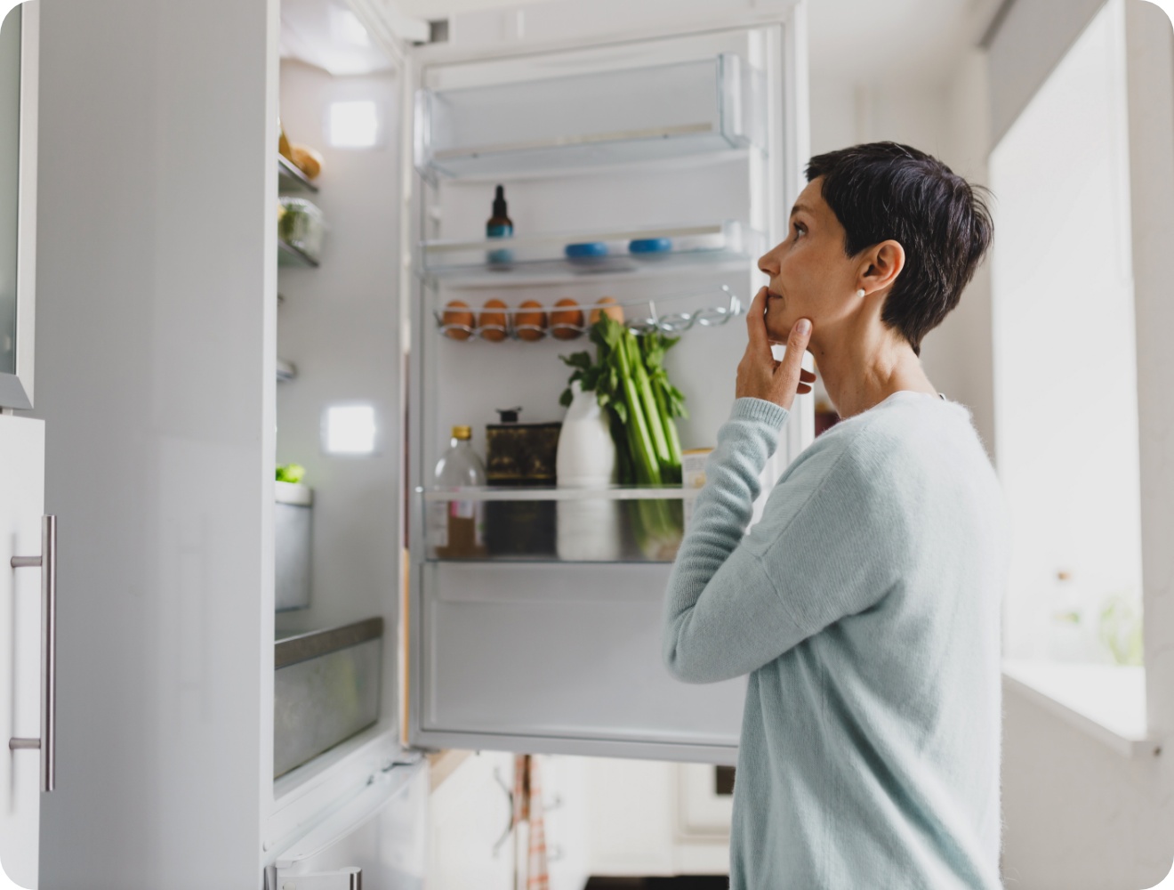 Frau in hellblauem Pullover schaut in einen offenem Kühlschrank gefüllt mit Eiern, Gemüse und Flaschen in einer hellen Küche.