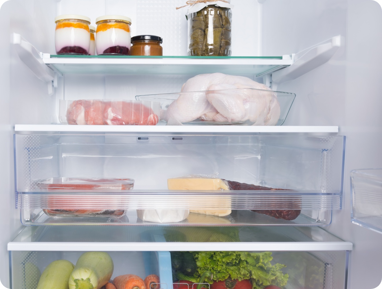 Kühlschrankfächer mit frischem Hühnchen, Fleisch, Käse, Joghurt, eingelegtem Gemüse und einem Gemüsefach mit Möhren und Salat.