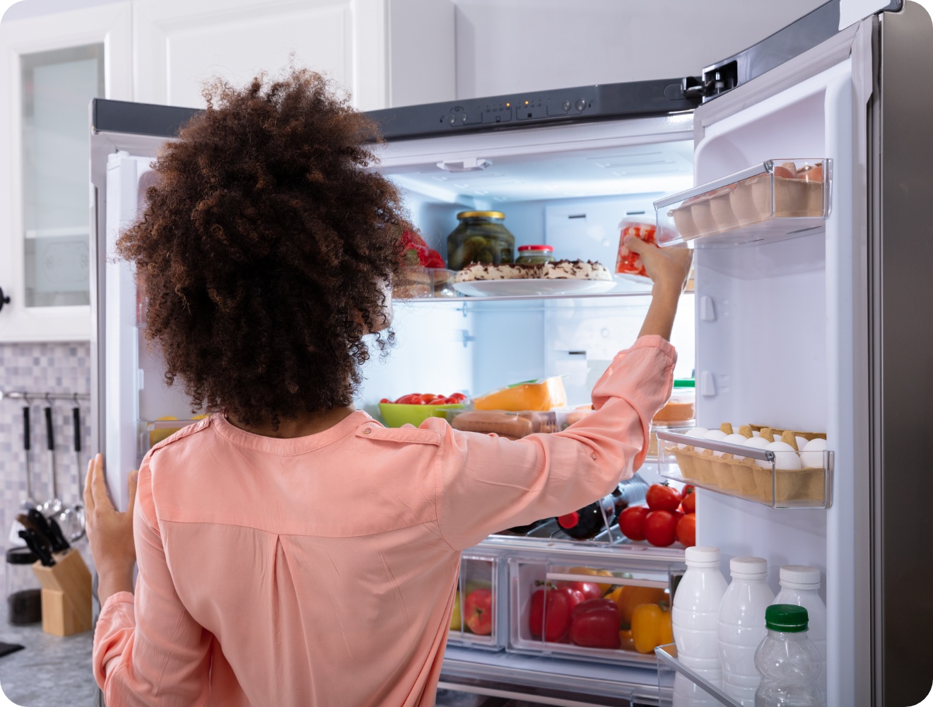 Frau in rosa Bluse steht vor offenem gefüllten Kühlschrank, greift nach einer Schale mit Lebensmitteln.