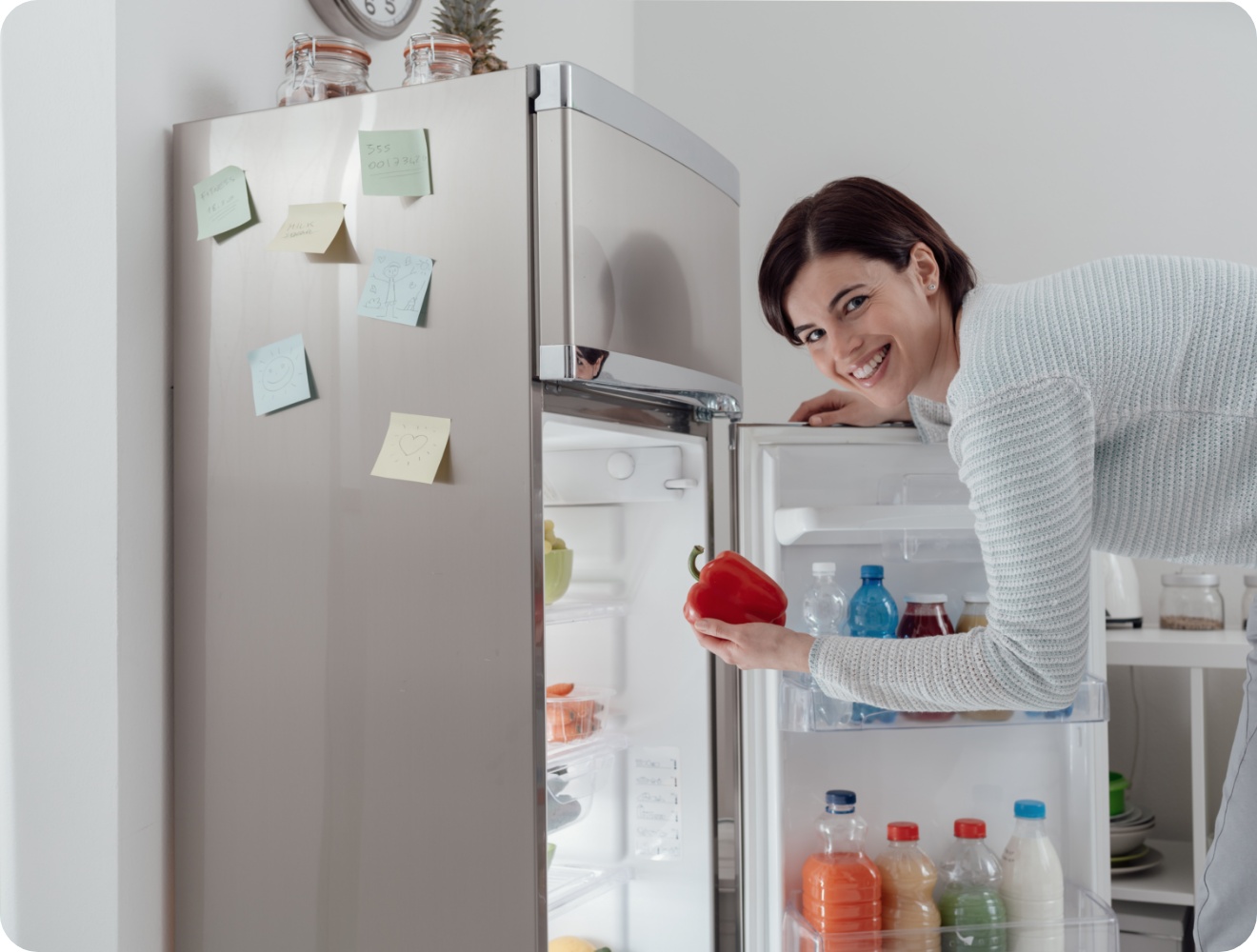 Lächelnde Frau holt eine rote Paprika aus einem offenen silbernen Kühlschrank.