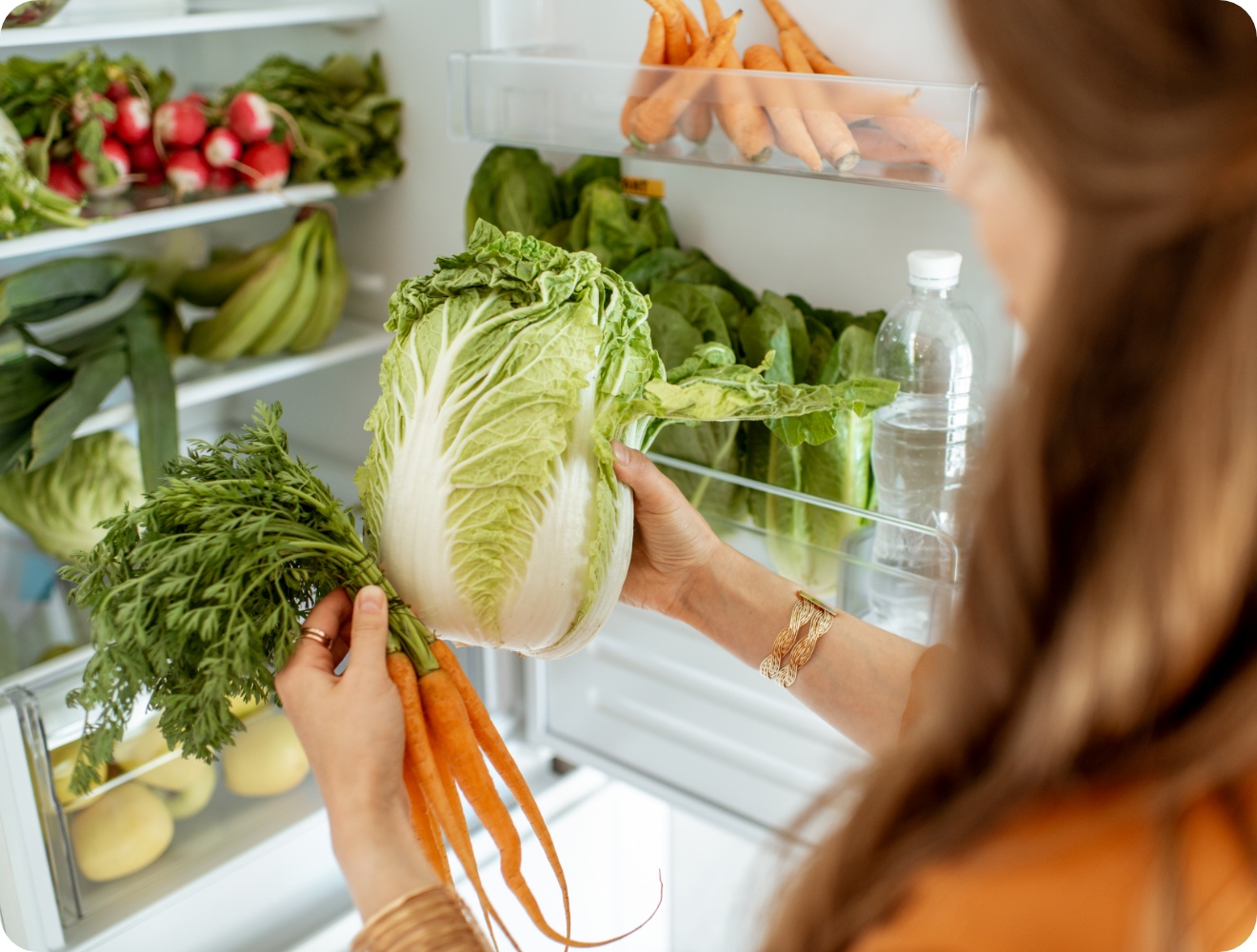 Frau räumt frisches Gemüse, darunter Chinakohl und Karotten, in einen gut gefüllten Kühlschrank mit gesunden Lebensmitteln ein.
