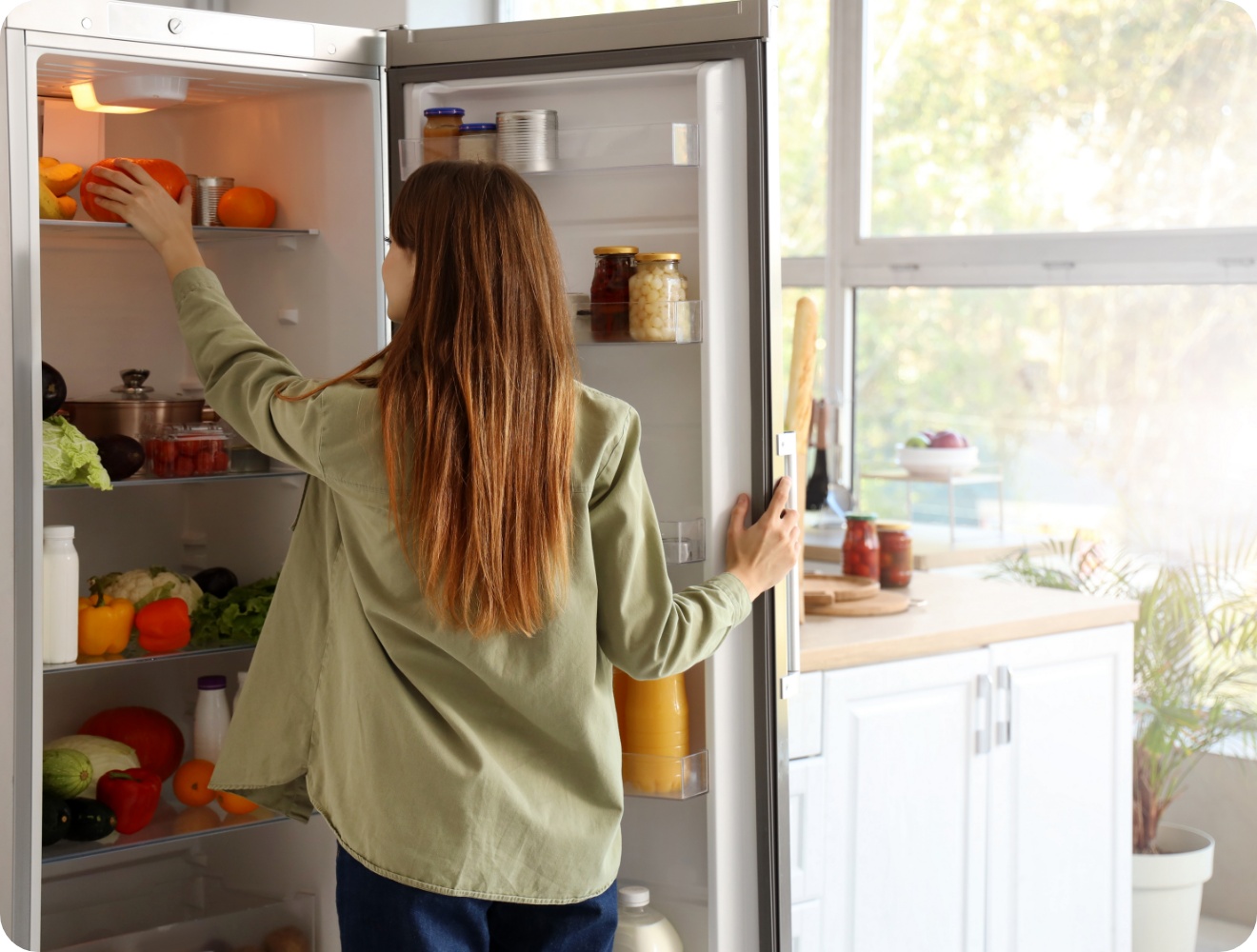 Frau mit langen Haaren räumt frische Paprika in einen gut gefüllten Kühlschrank in einer hellen, modernen Küche ein.
