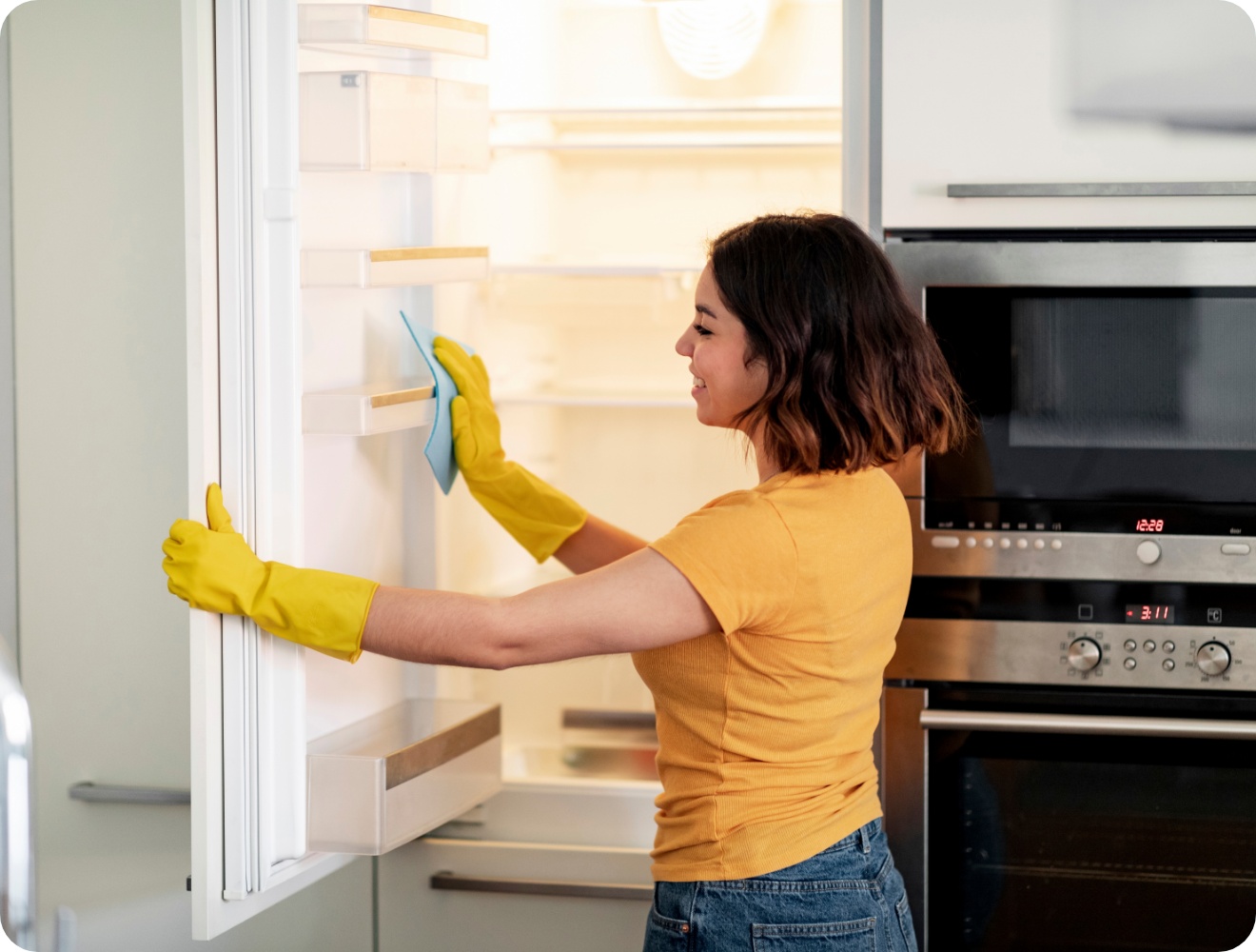 Frau mit gelben Gummihandschuhen reinigt die Tür eines leeren Kühlschranks mit einem blauen Tuch in einer modernen Küche.