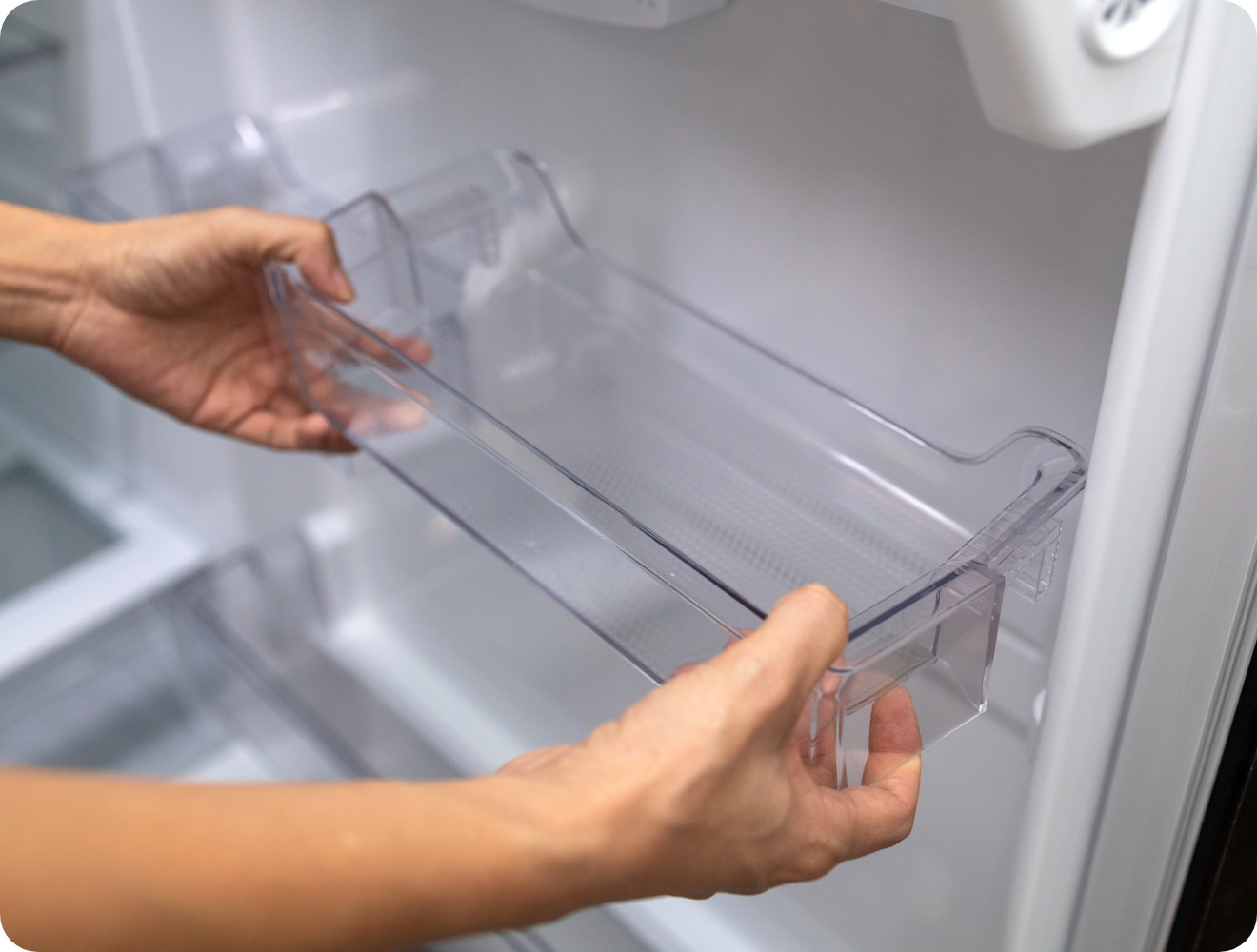 Nahaufnahme von Händen, die ein transparentes Kunststofffach in die Tür eines leeren Kühlschranks einsetzen oder es zur Reinigung entfernen.