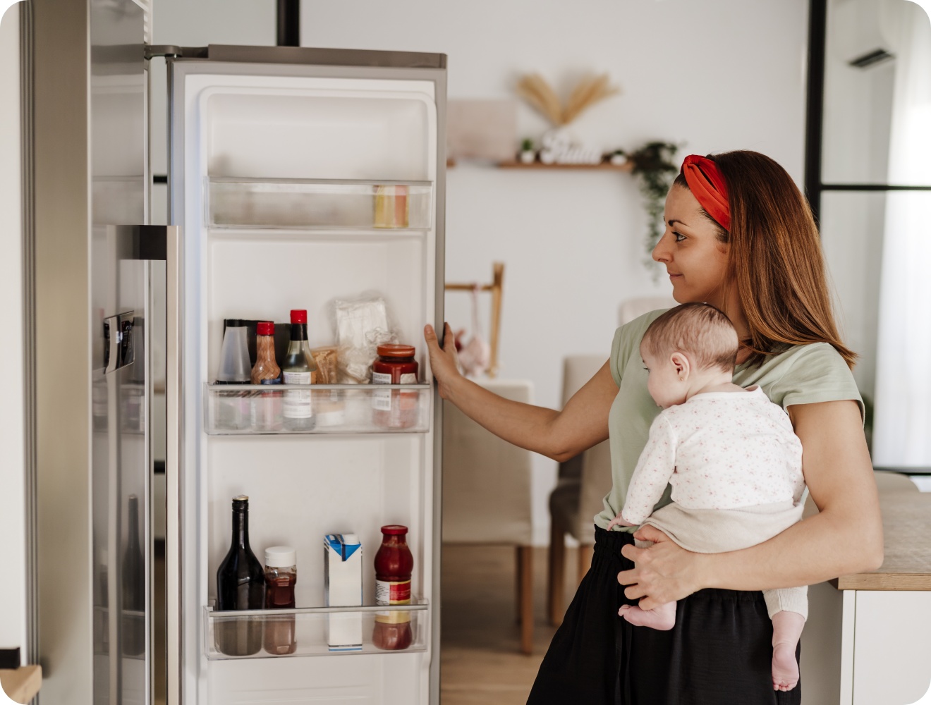 Mutter mit Baby auf dem Arm öffnet einen Kühlschrank mit verschiedenen Lebensmitteln und Saucen in einer modernen Küche.