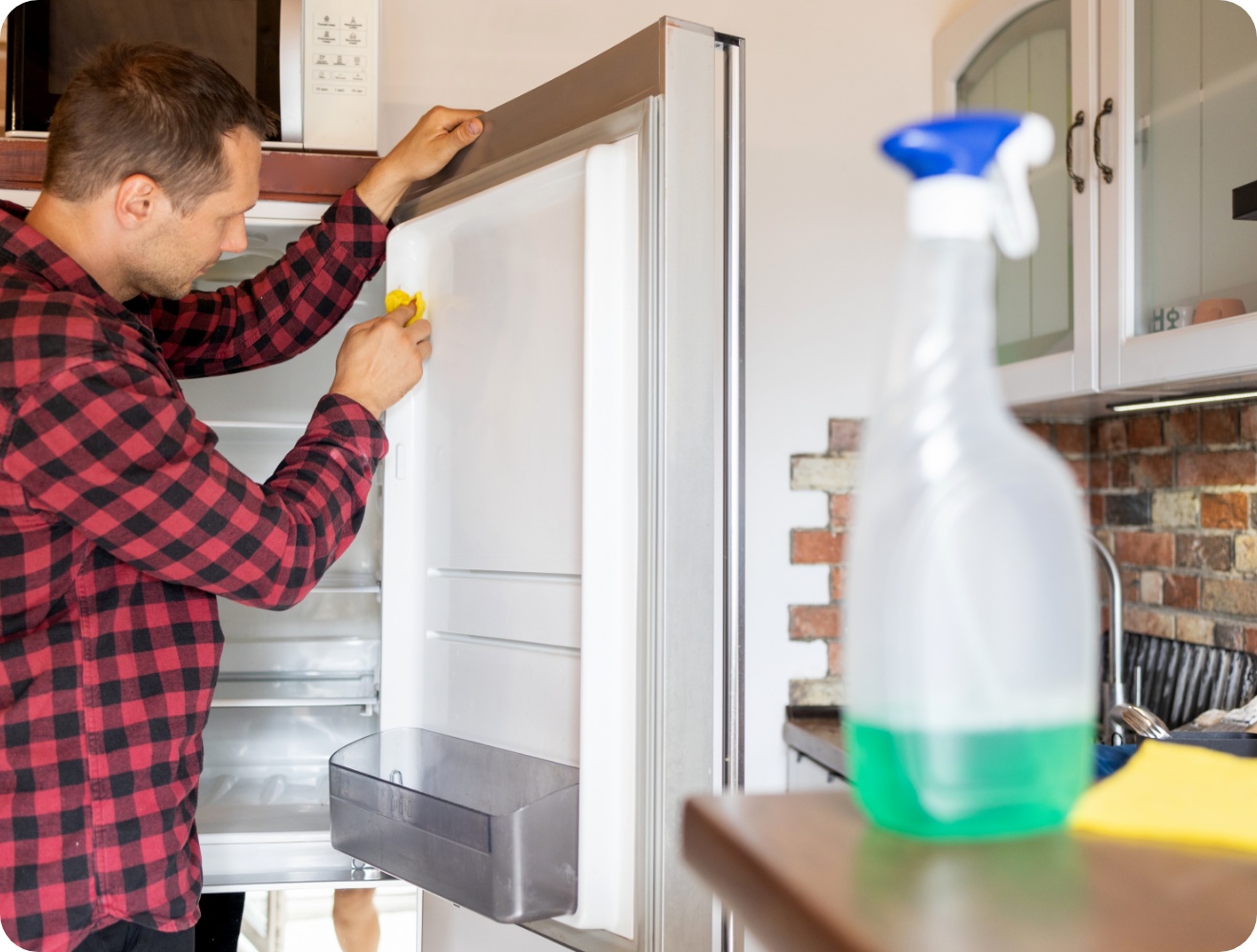 Mann in kariertem Hemd reinigt die Türdichtung eines offenen Kühlschranks mit einem gelben Tuch in einer rustikalen Küche.