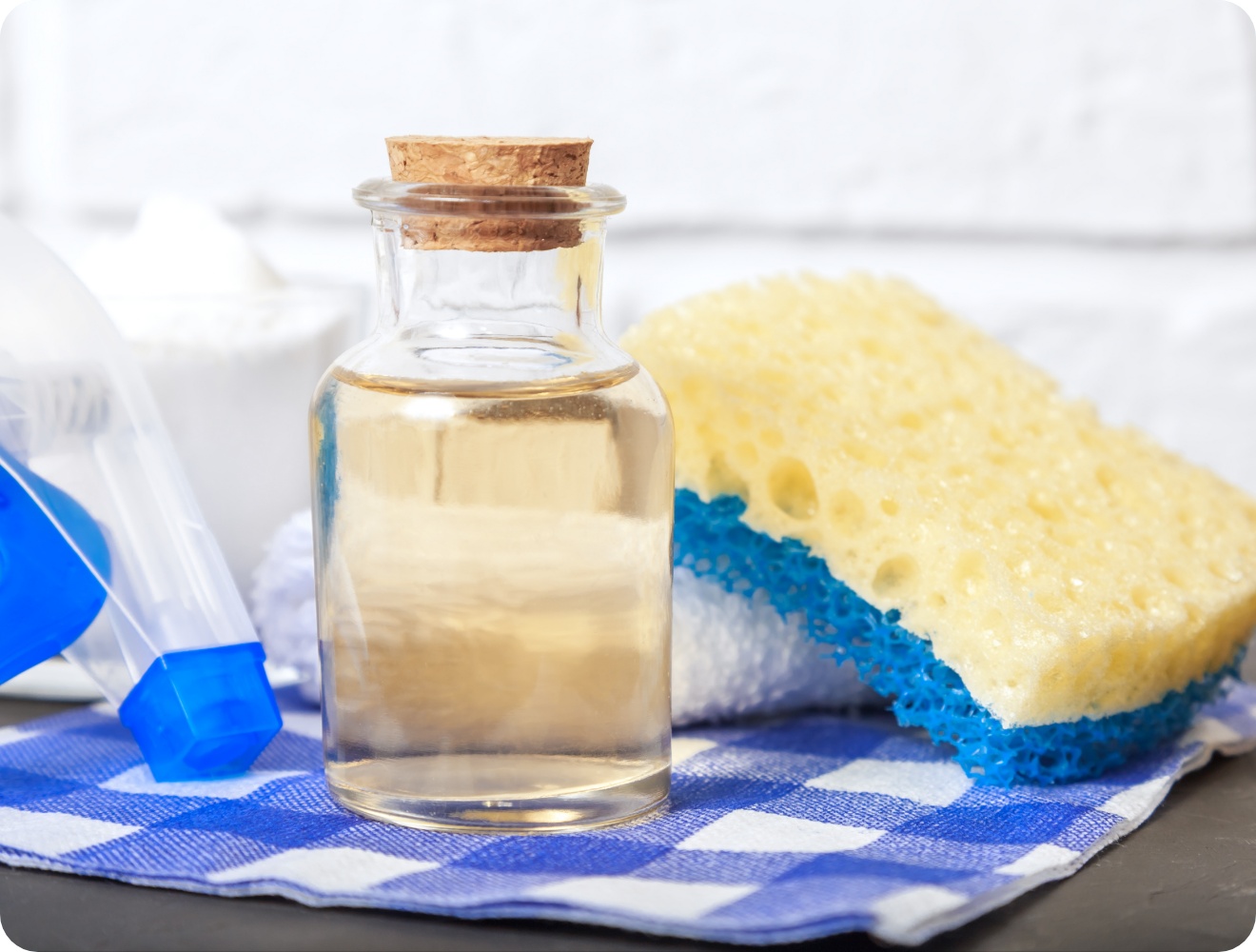 Glasflasche mit Essig, gelber Schwamm, Sprühflasche und Natron auf blau-weiß kariertem Tuch.