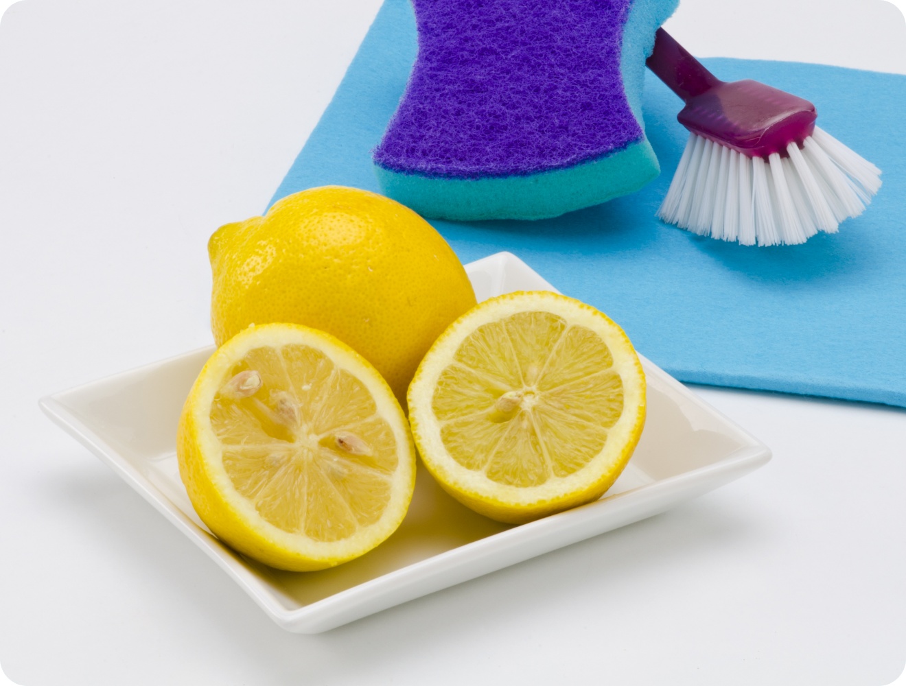 Zitronen auf weißem Teller, daneben blauer Schwamm, Reinigungsbürste und Tuch als natürliche Putzmittel.