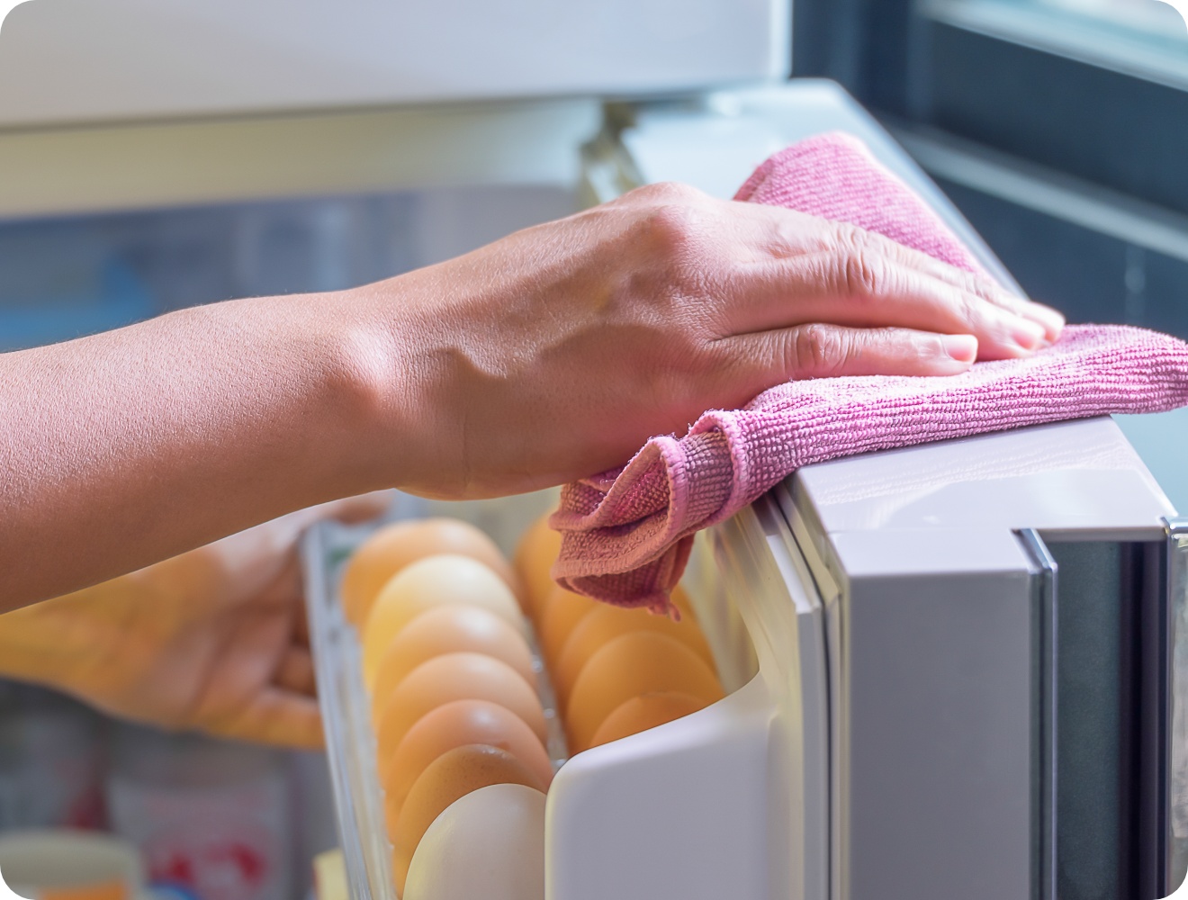 Eine Person wischt mit rosa Mikrofasertuch die Oberseite eines Kühlschranks neben einem Eierfach sauber.
