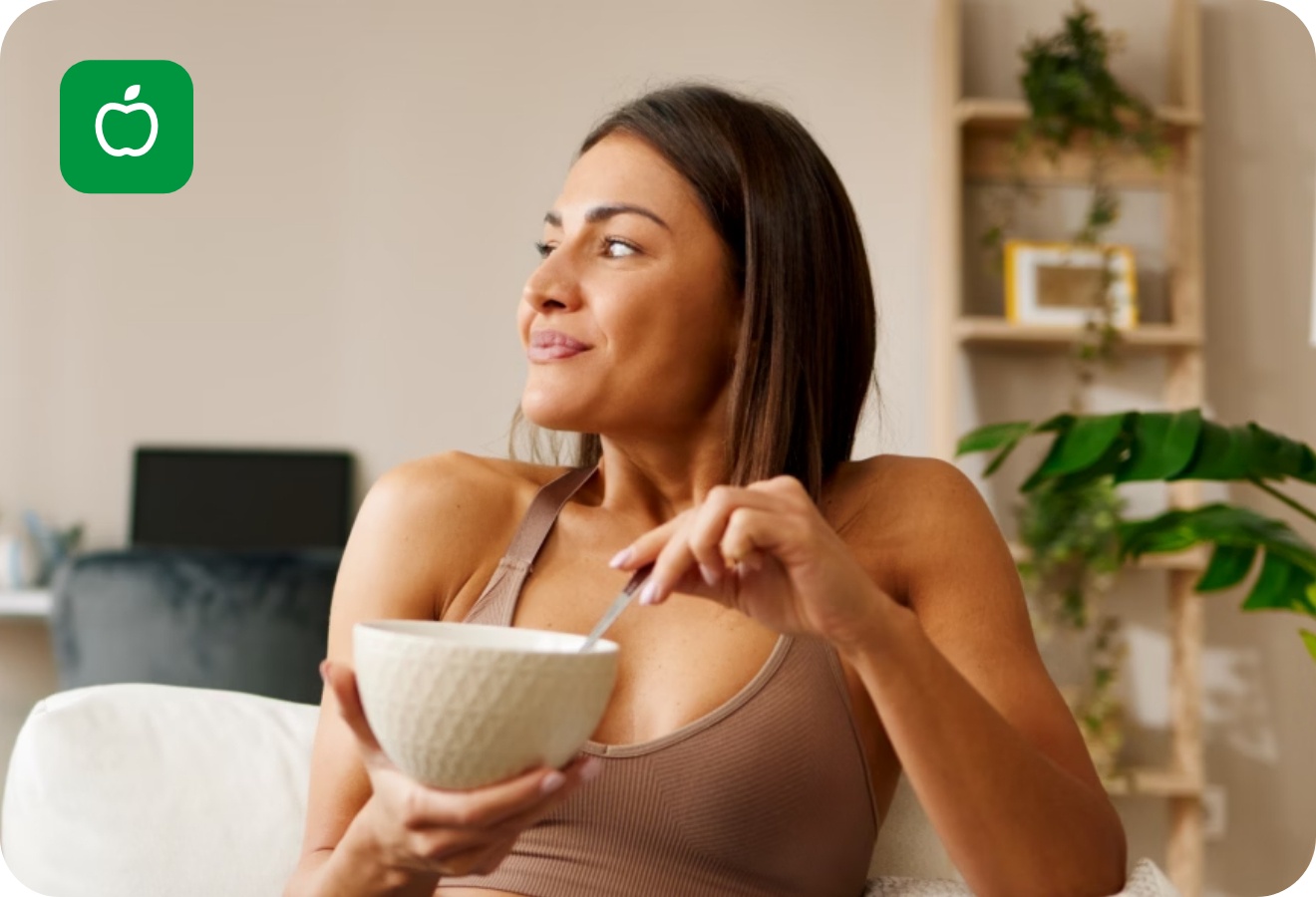 Frau in Sportkleidung sitzt auf einem weißen Sofa und isst lächelnd eine Bowl.