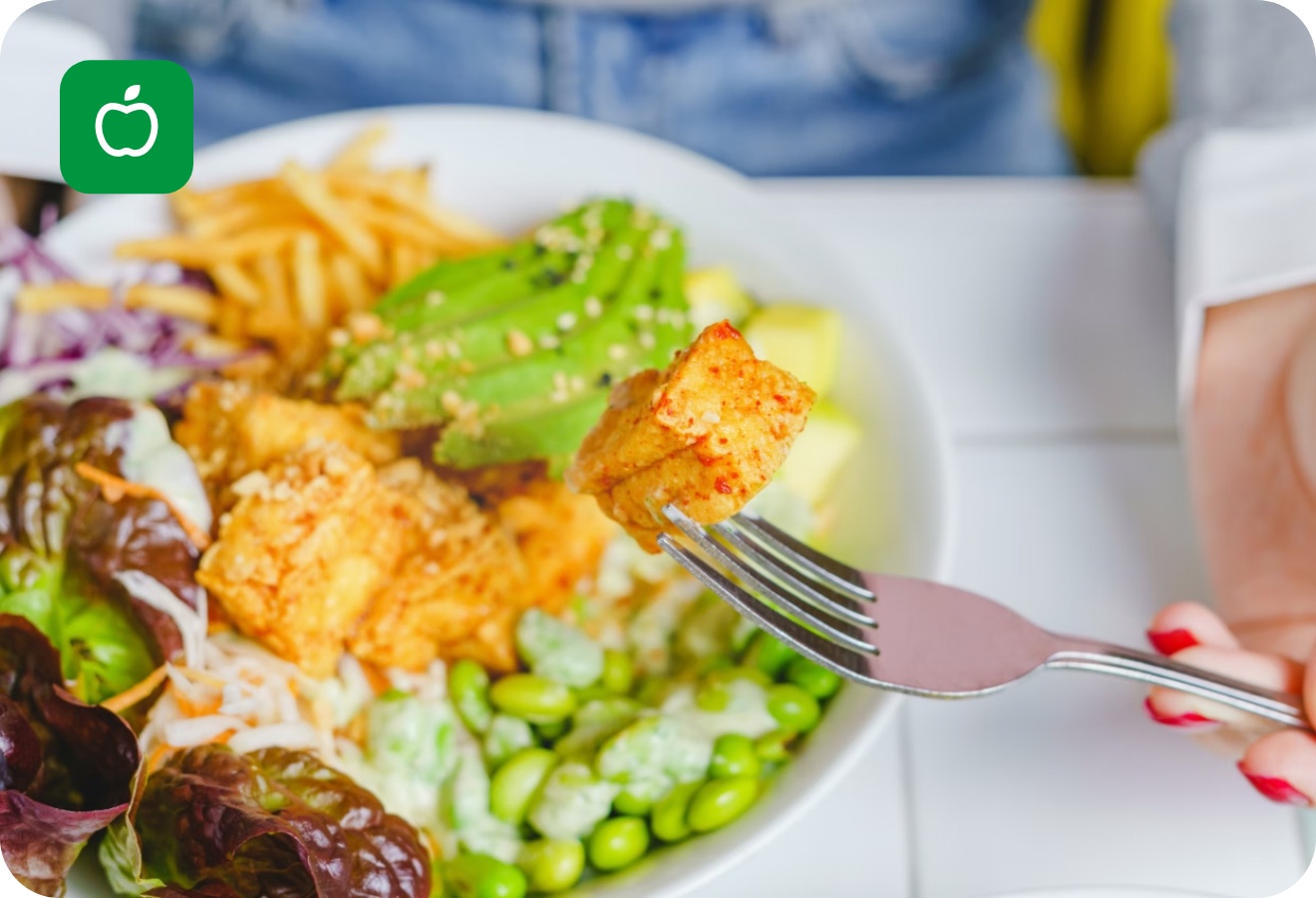 Gabel mit Tofu-Stück über einer farbenfrohen Salatschüssel mit Avocado, Edamame, Pommes und Blattsalat.