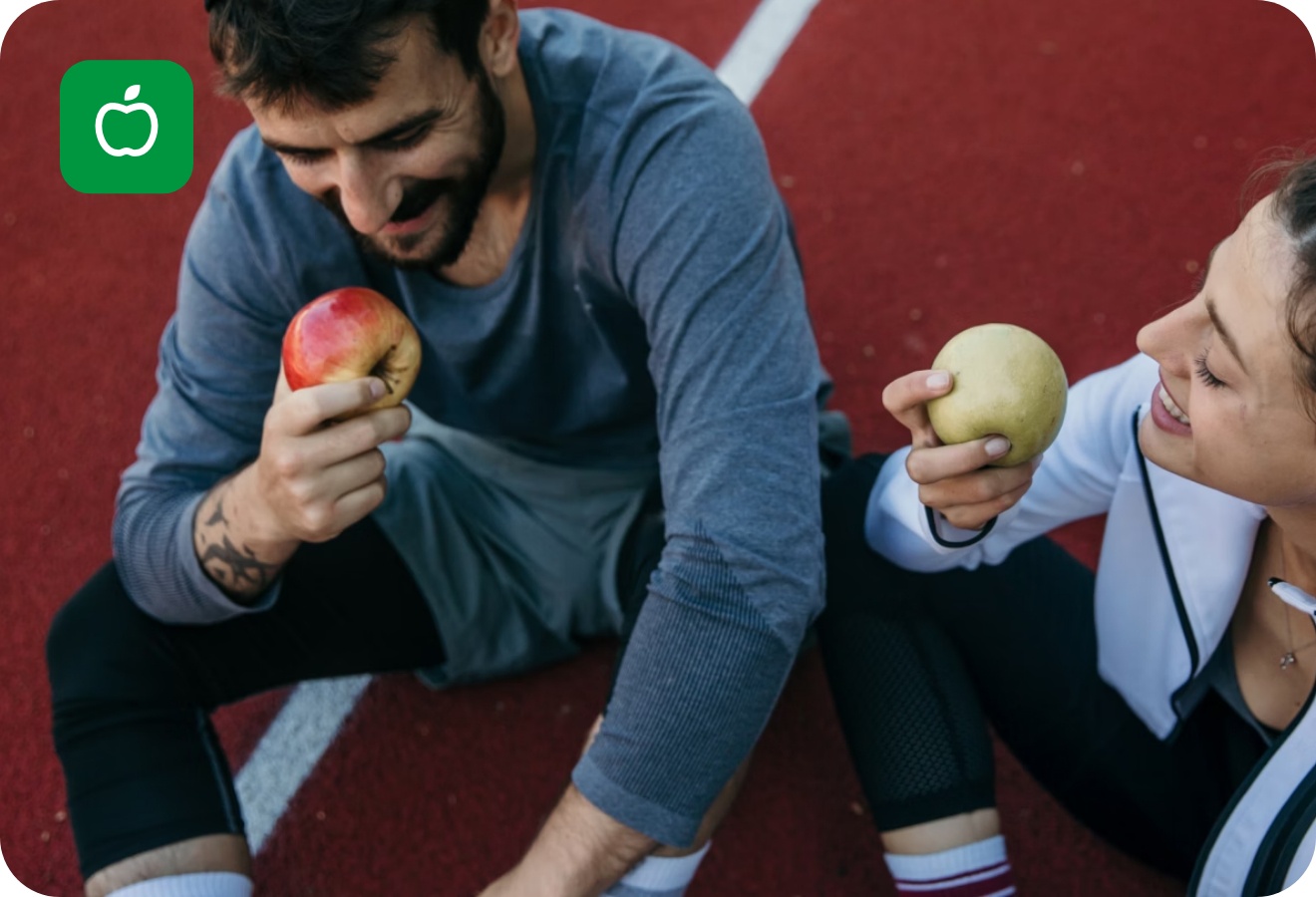 Zwei Personen in Sportkleidung sitzen auf einer Laufbahn und essen lächelnd Äpfel nach einem Training.