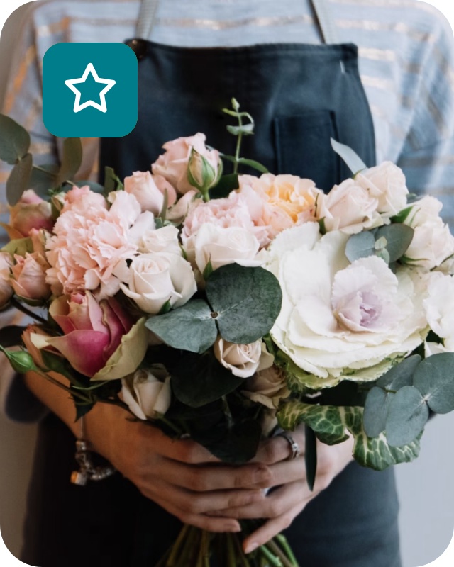 Floristin mit dunkelgrüner Schürze hält Blumenstrauß aus weißen und hellrosafarbenen Blumen.