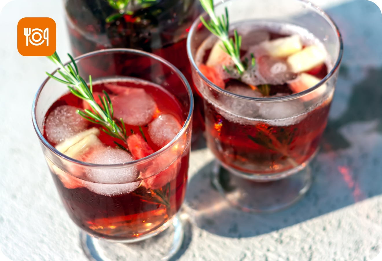 Zwei Gläser roter Sommerdrink mit Eiswürfeln, Rosmarin, Erdbeeren und Ingwer auf einem Tisch in der Sonne.