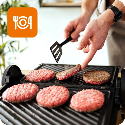 Eine Person grillt Burger-Patties auf einem Kontaktgrill. Senf, Ketchup und geschnittene Zwiebeln liegen bereit.