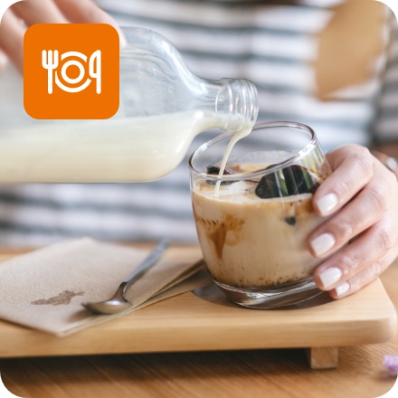 Person gießt Milch aus einer Glasflasche in ein Glas mit Eiskaffee auf einem Holztablett.