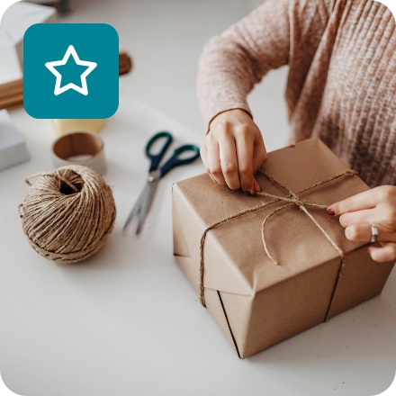 Frau verpackt Geschenk in braunem Papier mit Juteschnur, umgeben von Verpackungsmaterialien.