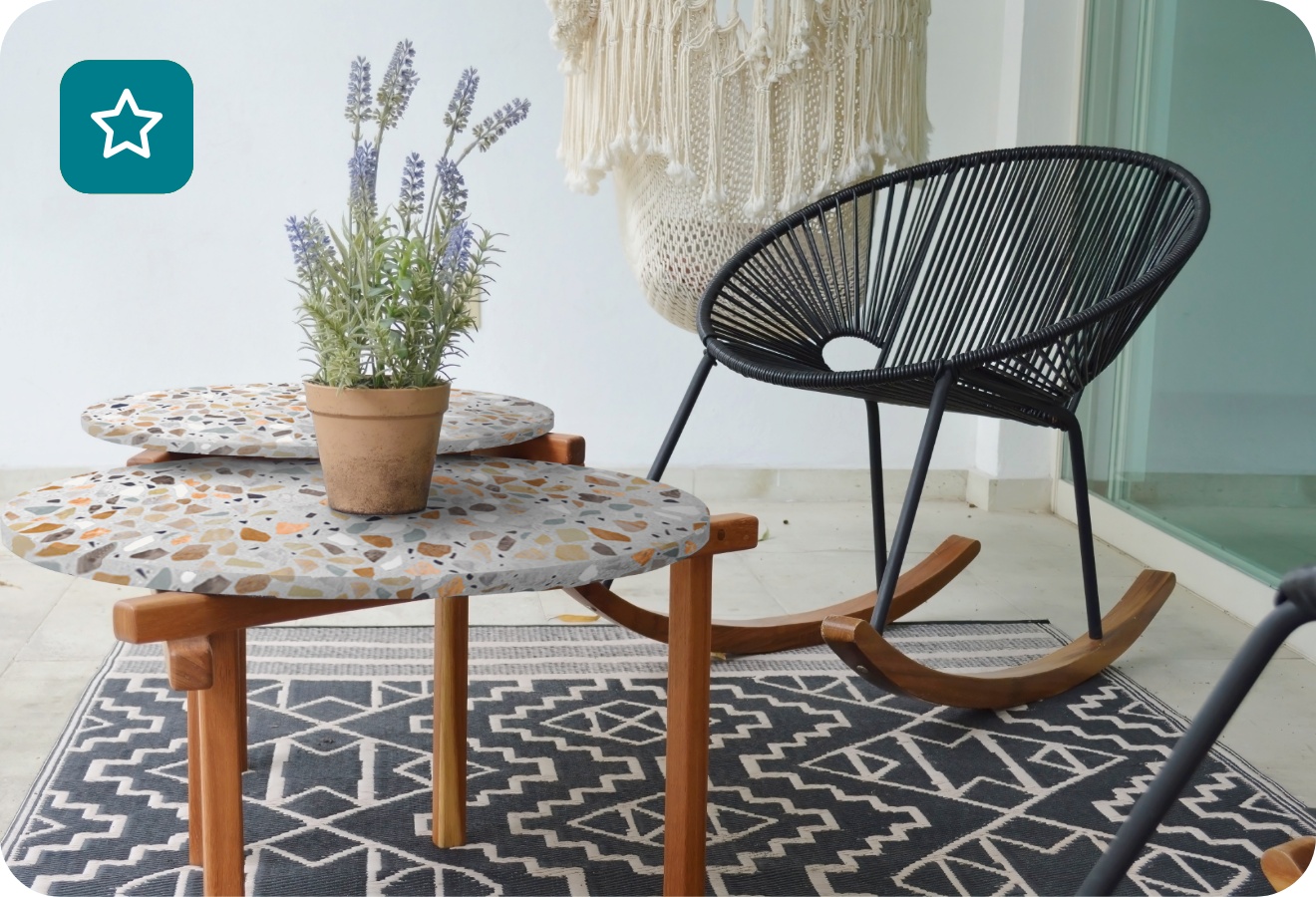 Zwei unterschiedlich große Tischplatten in Terrazzo-Optik mit Tischbeinen aus Holz zusammen mit runden Rattan-Schaukelstühle auf einem Balkon.