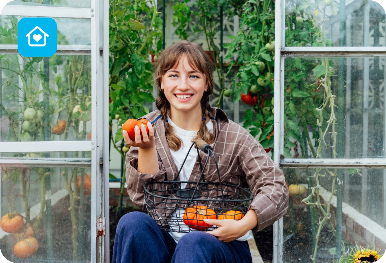 Frau sitzt im Gewächshaus und hält eine frisch geerntete Tomate und einen Korb mit weiteren Tomaten.