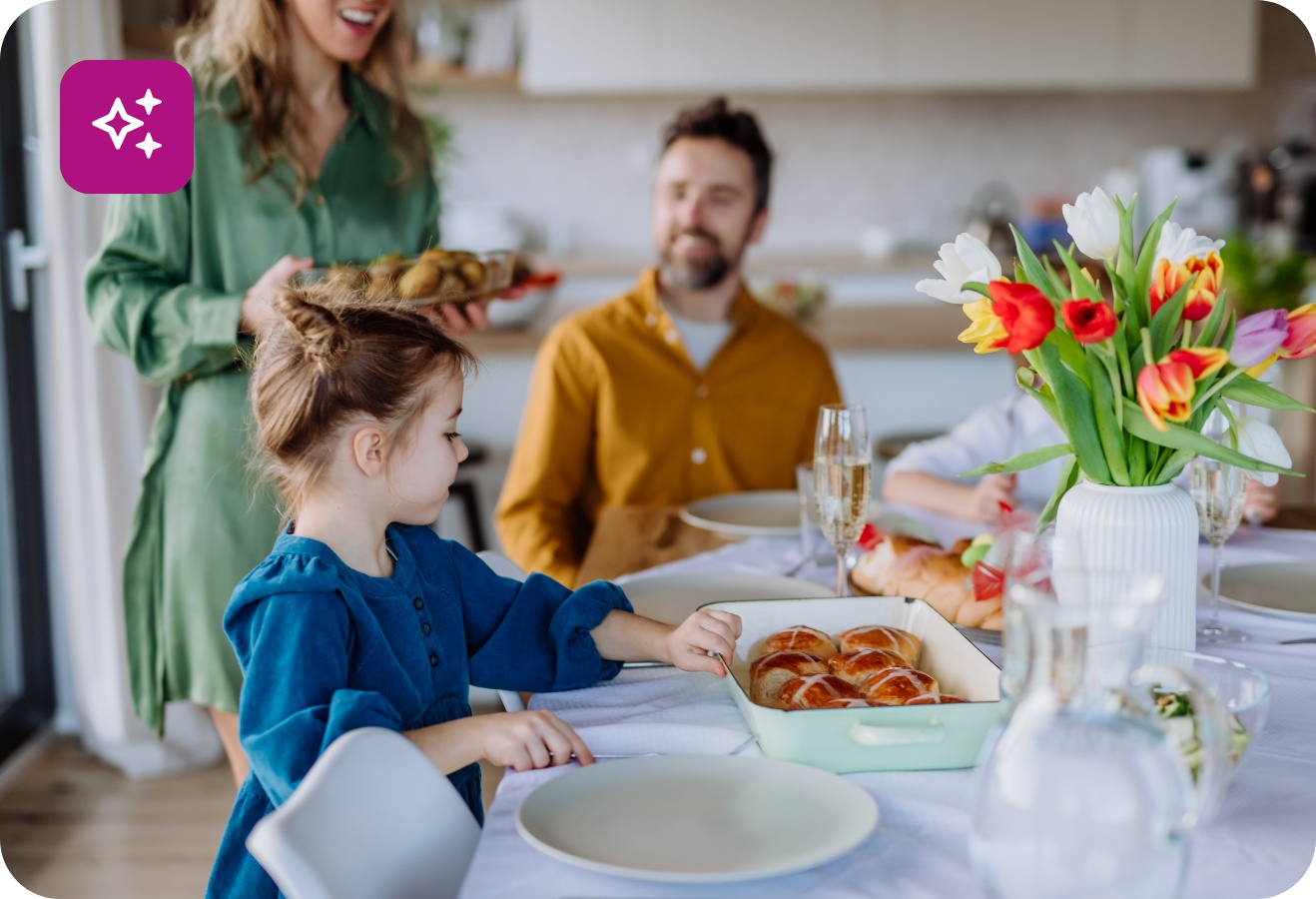 Eine Familie sitzt bei einem Osterbrunch beisammen. Auf dem Tisch stehen Tulpen und Ostergebäck wie süße Brötchen und ein Hefezopf.