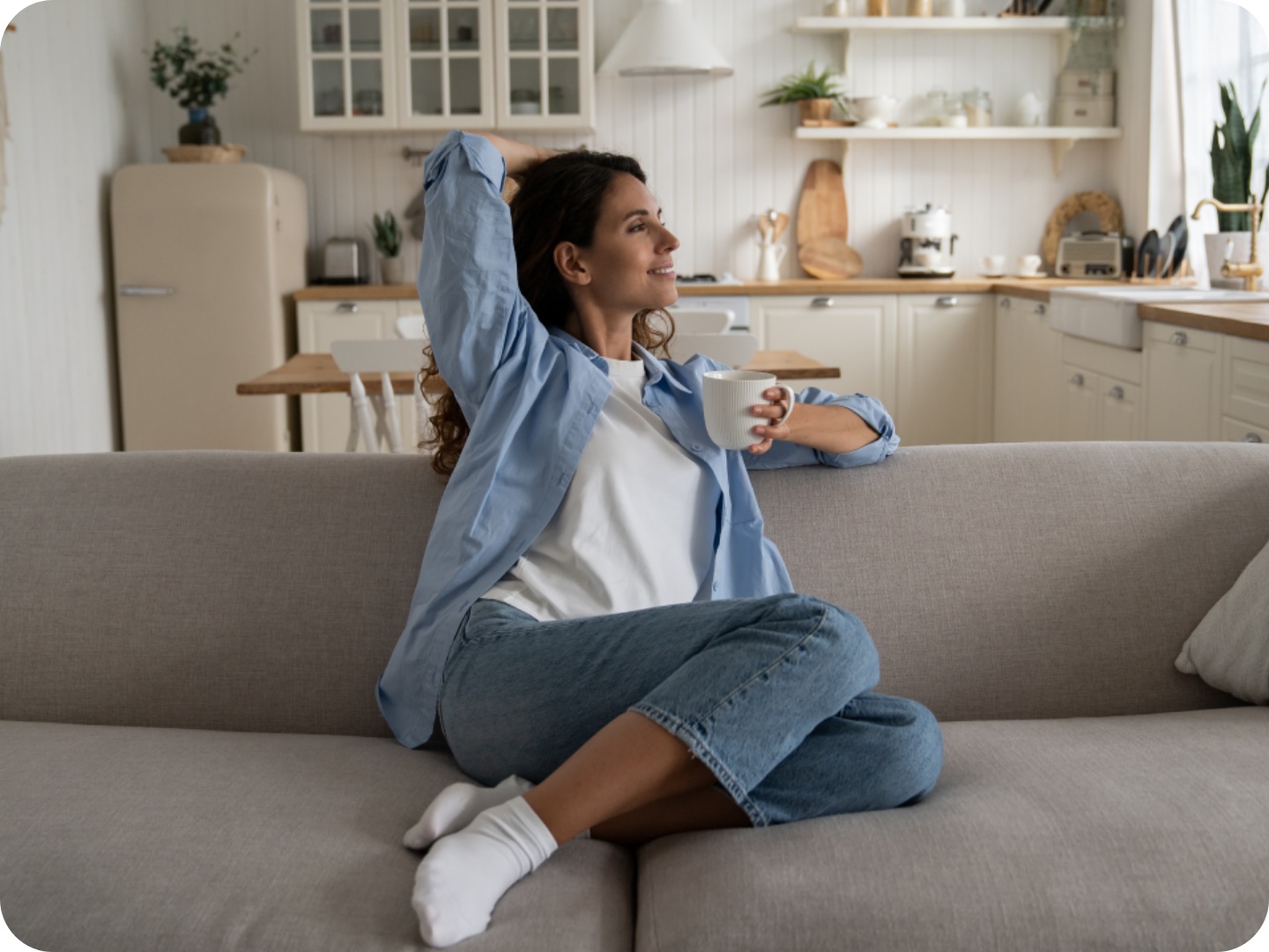 Frau sitzt entspannt auf einem Sofa mit einer Tasse Kaffee in einer hellen aufgeräumten Wohnküche.
