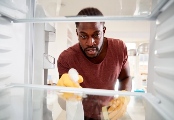 Eine Mann sprüht Reinigungsmittel in den ausgeräumten Kühlschrank.