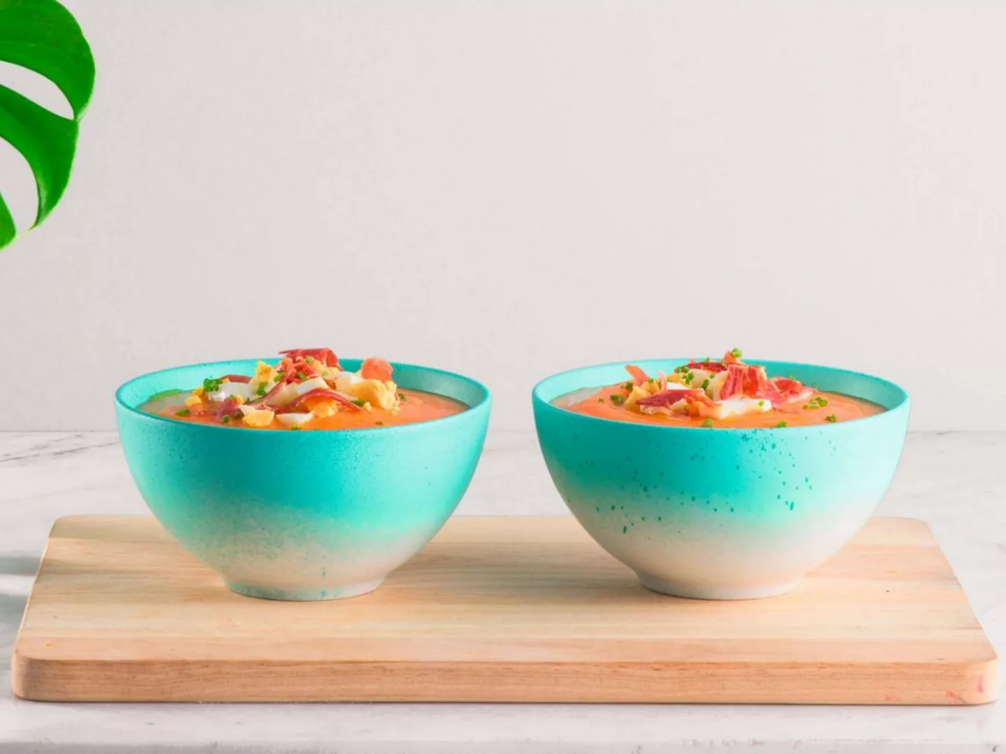 Dos boles de salmorejo servidos en cuencos turquesa, decorados con huevo duro picado, jamón serrano y cebollino, sobre una tabla de madera clara.
