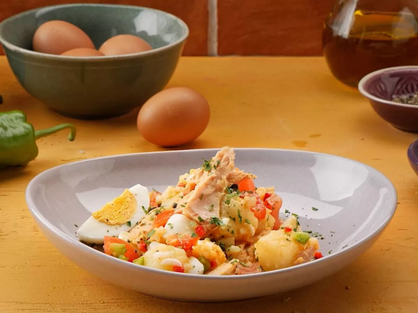 Ensalada campera con patata, huevo duro, atún y verduras sobre plato gris, con huevos frescos y aceite al fondo