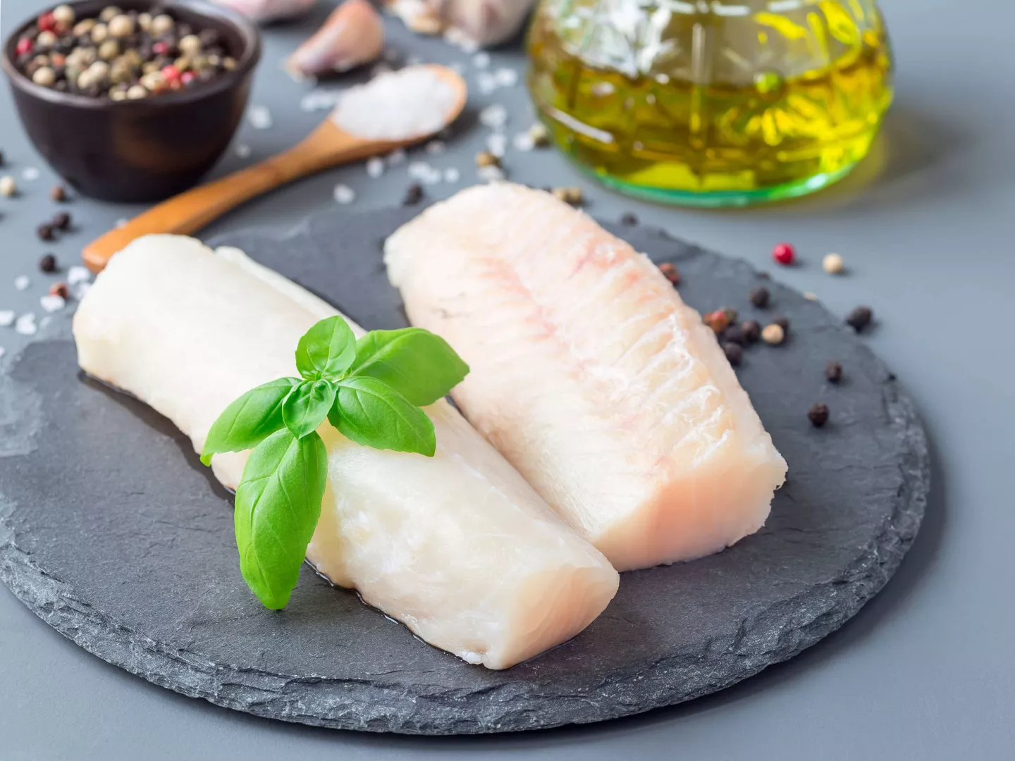 Filetes crudos de merluza fresca sobre plato de pizarra con hoja de albahaca, listos para cocinar a la plancha.