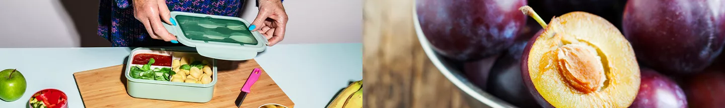 Eine Schüssel mit Pflaumen, eine aufgeschnittene mit Kern im Fokus, links wird eine Lunchbox geschlossen.