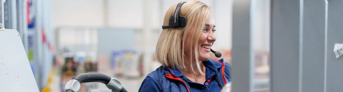 Mitarbeiterin im Logistikbereich trägt ein Headset und lächelt, neben ihr ist das Lenkrad eines Fahrzeugs, bzw. eines Staplers zu sehen. 