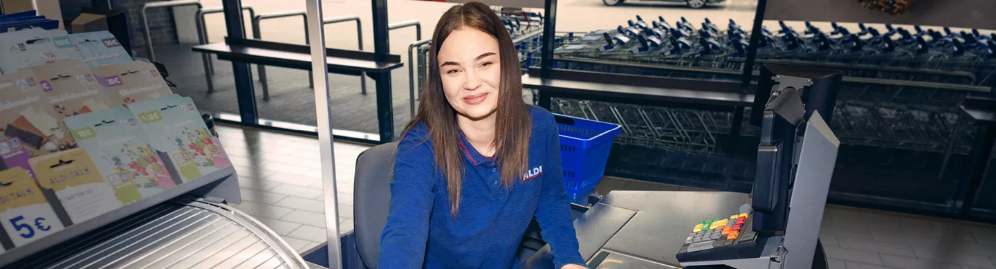 Junge Frau im blauen Shirt sitzt an der Kasse und blickt in die Kamera; im Hintergrund sind Einkaufswagen und der Ausgangsbereich zu sehen.