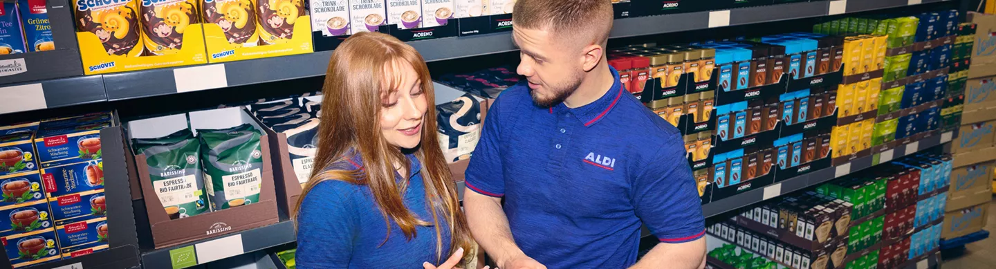 Zwei Personen in Arbeitskleidung stehen im Markt vor einem Regal mit Kaffee- und Getränkepackungen und sprechen miteinander.