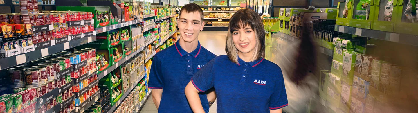 Zwei junge Mitarbeitende in ALDI Poloshirts stehen lächelnd im Supermarktgang zwischen vollen Regalen.