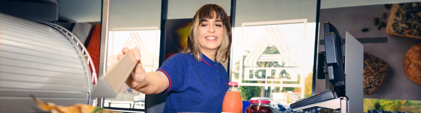 ALDI Mitarbeiterin im blauen Shirt an der Kasse reicht einer Kundschaft den Kasseneinleger. Im Vordergrund stehen Getränke und Waren am Kassenband.