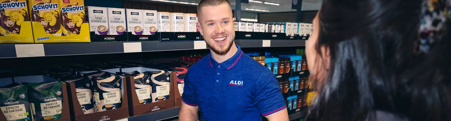 Ein freundlicher Mitarbeiter im blauen ALDI-Poloshirt steht lächelnd in einem Gang vor Regalen mit Kaffee- und Schokoladenprodukten und unterhält sich mit einer Kundin, die mit dem Rücken zur Kamera steht.