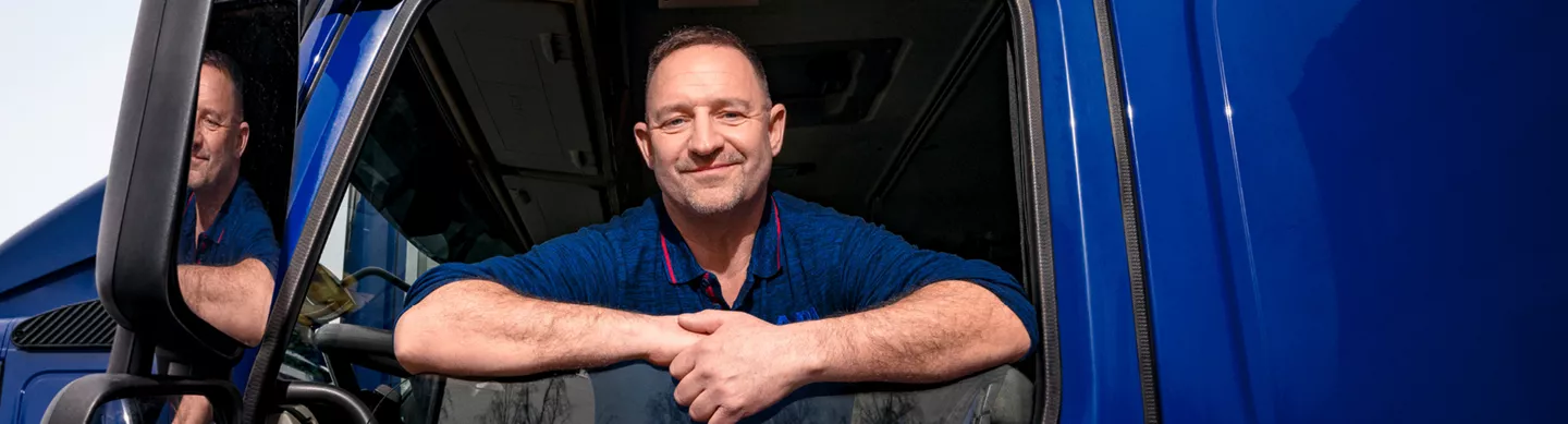 Ein Mann in Berufskleidung lehnt sich aus dem Fenster der Fahrerkabine eines modernen LKW. Er lächelt selbstbewusst in die Kamera.
